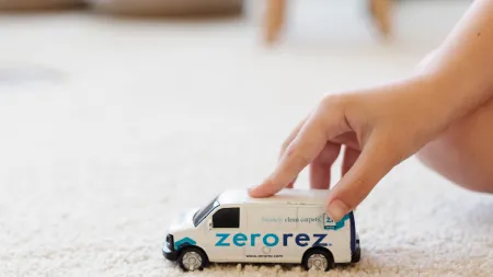 Child's hand playing with a toy Zerorez van on a clean beige carpet in a bright room.