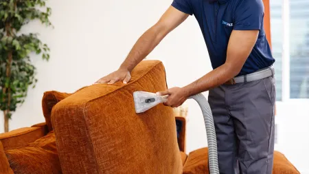 Man in zerorez uniform professionally cleaning orange sofa upholstery with steam cleaning in bright living room