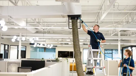 Technicians servicing HVAC ductwork in a modern office space using equipment and ladder under ceiling vents.