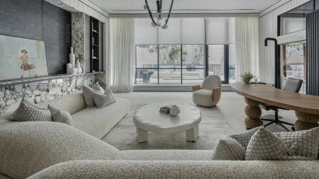 Modern living room with curved sofas, irregular white coffee table, wooden desk, and large windows with natural light.